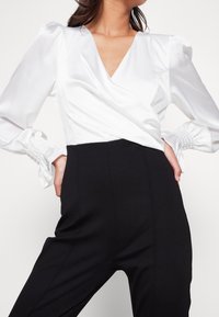 Vit satinwrap-topp med puffärmar och smockade manschetter, ihopparad med högmidjade svarta byxor. Slät textur och figursydd design.