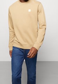 Felpa beige con colletto a girocollo, polsini e orlo a costine, caratterizzata da un piccolo logo quadrato sul lato sinistro del petto. Abbinata a jeans blu.