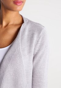 Cardigan tricoté gris clair avec un tissu texturé et un design ouvert à l'avant, comportant un tissu de poids moyen et une coupe décontractée.