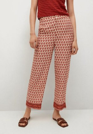 Kobieta nosząca czerwoną teksturowaną bluzkę bez rękawów oraz szerokie spodnie z wysokim stanem, z czerwonym wzorem liści i detalem na brzegach, w połączeniu z brązowymi sandałami typu slide.