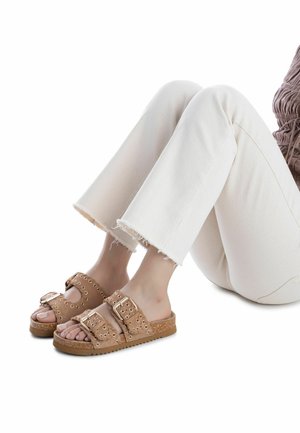 Sandalias tipo slide de gamuza marrón con dos tiras anchas con hebillas, diseño perforado y suela de corcho. Acompañadas de jeans blancos recortados con dobladillo deshilachado.