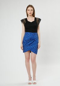 Zwarte top met kanten fladdermouwen, gecombineerd met een blauwe geprinte wikkelrok met drapering. Model draagt witte open teen hakken.