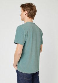 Jonge man met kort bruin haar, gekleed in een geribbelde saliegroene T-shirt en donkerblauwe jeans, staand met zijn rug naar ons toe en met zijn handen in zijn zakken.