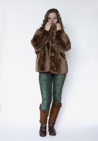 Bruine nepbonten jas met grote knopen, groene textuurrijke legging en bruine laarzen tot net onder de knie. Eenvoudig ontwerp, warme materialen en contrasterende kleuren.