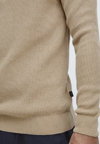 Beige strikkegenser med teksturert mønster, ribbede mansjetter og kant. Har et lite svart merke på siden. Nærbilde av stoffet på en arm.