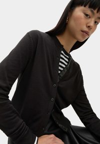 Cardigan noir avec fermeture à bouton, poignets et ourlet côtelés. Porté par-dessus une chemise à rayures noires et blanches, mettant en avant un look superposé.