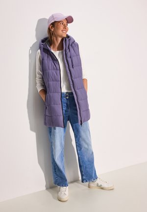 Gilet matelassé de couleur violet clair avec une capuche, associé à un t-shirt à manches longues blanc et un jean large bleu clair, complété par des baskets blanches.