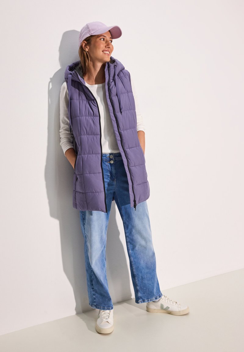 Puffweste in Helllila mit Kapuze, kombiniert mit einem weißen Langarmshirt und hellblauen weiten Jeans, ergänzt durch weiße Sneakers.