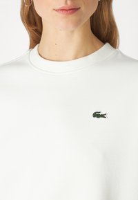 Felpa bianca con texture e scollatura rotonda. Presenta un piccolo logo di coccodrillo ricamato in verde sulla parte sinistra del petto.