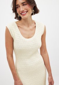 Neulemekko off-white-värissä, jossa on avara pääntie ja tyköistuva siluetti. Teksturoitu ribbikuvio, lyhyet hihat ja pehmeä, joustava kangas.