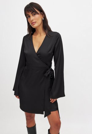 Abito nero a tubino realizzato in tessuto liscio, con maniche lunghe, scollatura a V, dettaglio in vita con cintura annodata e uno spacco alto su un lato.