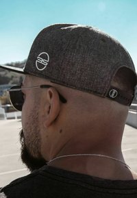 Graue Stoffmütze mit flachem Schirm, auf der Seite mit einem runden Logo versehen. Dunkle Sonnenbrille und kurze Haare sind auf der Rückseite sichtbar.