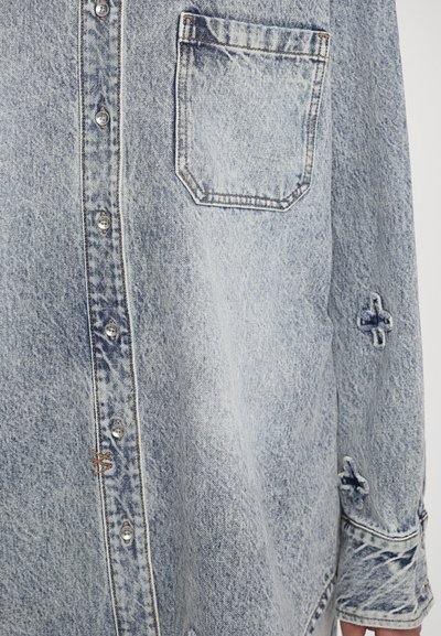 Šviesiai mėlynas džinsinis marškinis su kišenėle, mygtukų uždarymu ir tekstūruota medžiaga. Rankovėse yra siuvinėti gėlių akcentai.