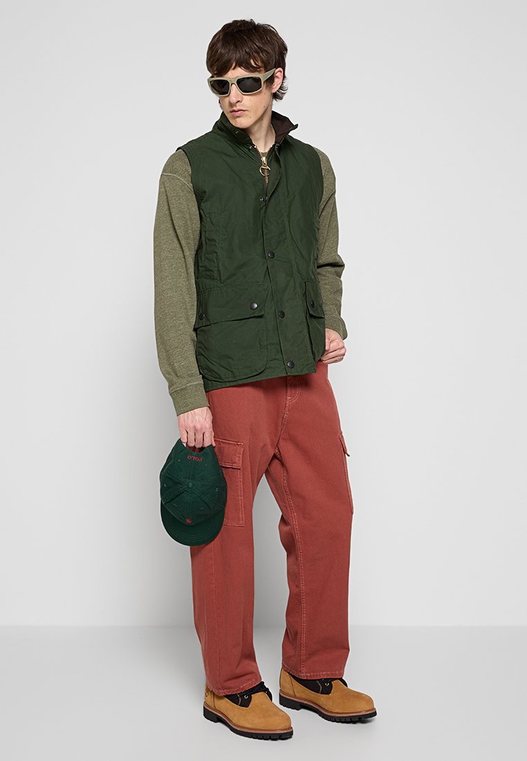 Mann trägt eine grüne Steppweste über einem olivfarbenen Pullover, rostfarbene Cargo-Hosen, sandfarbene Stiefel, hält eine grüne Kappe in der Hand und trägt Sonnenbrillen vor einem grauen Hintergrund.