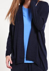 Námořnický kardigan vyrobený z měkkého pleteného materiálu, s otevřeným předním dílem a žebrovanou texturou na rukávech, nošený přes světle modré tričko.