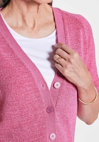 Heller rosa Cardigan mit V-Ausschnitt-Design, ausgestattet mit drei rosa Knöpfen. Über einem weißen Hemd getragen, das einen texturierten Stoff zeigt.