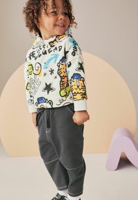 Farverig hættejakke med tegneserie dyremotiver, hvid med multikolorede designs. Grå sweats, løs pasform, tekstureret stof, sorte sko.