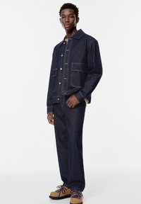 Giubbotto in denim scuro con cuciture in vista e tasche frontali, abbinato a jeans scuri e scarpe color nocciola con lacci blu.