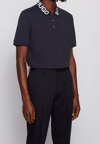 Námořnická polo košile s texturovanou tkaninou a bílým značkovým límečkem. Vypadající s tmavými kalhotami, které zdůrazňují čistý a ležérní vzhled.