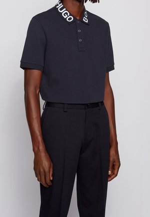 Marineblå polo t-shirt med tekstureret stof og en hvid mærket krave. Kombineret med mørke bukser, hvilket fremhæver et rent og afslappet udseende.