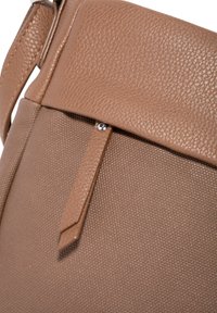 Tasche aus braunem Leder mit strukturiertem Oberflächenfinish, ausgestattet mit einem Klappenverschluss und einem metallischen Akzent. Der Riemen und das Design sind minimalistisch.