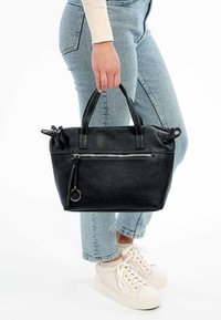 Svart strukturerad läderhandväska med dubbla handtag, frontficka med dragkedja och cirkulär detalj i metall. Bärs av en person i avslappnade jeans.