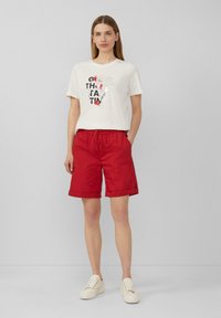 Witte t-shirt met zwart en rode grafische tekst, gecombineerd met rode shorts met een elastische tailleband en omgeslagen zomen. Witte sneakers.