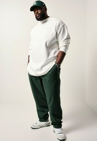 Witte shirt met lange mouwen, groene broek en witte sneakers met groene accenten. De persoon draagt een groene pet met een wit logo. Casual look.