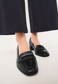Mocassins en cuir noir avec un bout carré, dotés d'une bande d'accent noire brillante. Semelle à talon, texture lisse et design minimaliste.