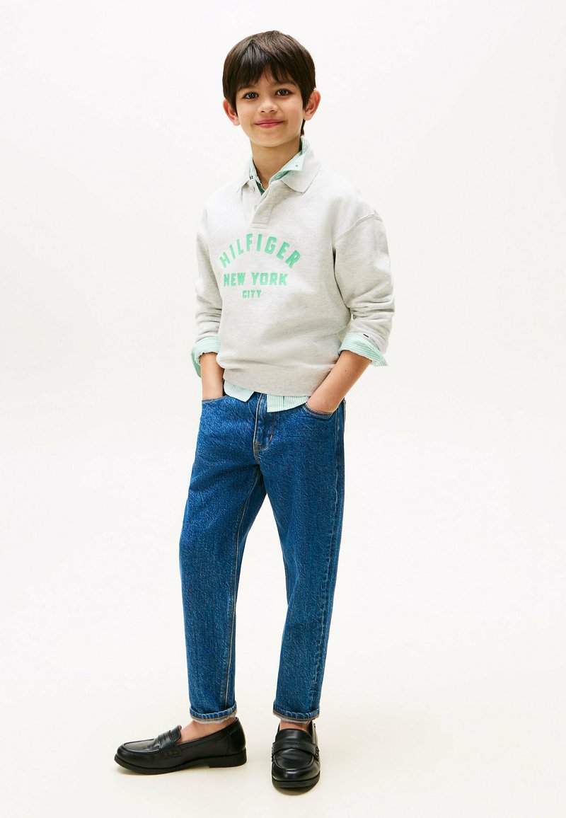 Felpa grigia chiara con la scritta "HILFIGER NEW YORK CITY" in testo verde, abbinata a jeans blu texture e mocassini neri.