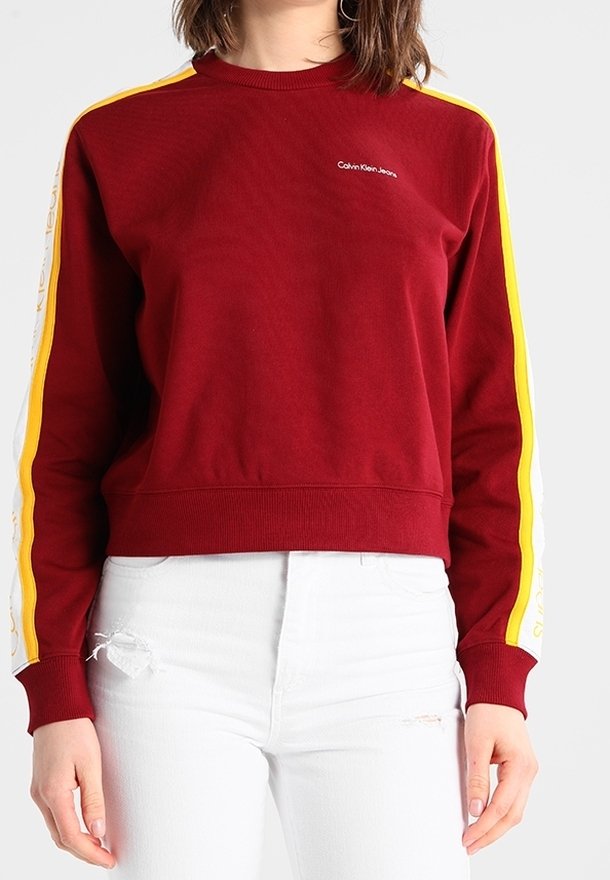 Femme portant un sweat-shirt rouge Calvin Klein avec des rayures jaunes et blanches sur les manches, associé à un jean blanc déchiré.