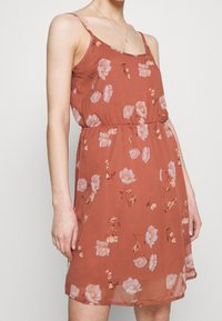 Abito color ruggine con motivo floreale, realizzato in tessuto leggero. Presenta spalline sottili e una vita elastica per una vestibilità comoda.
