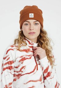 Braune gerippte Mütze mit Logo-Tag, getragen mit einem weißen und roten gemusterten Oberteil mit Reißverschlusskragen. Das Haar ist wellig und schulterlang.