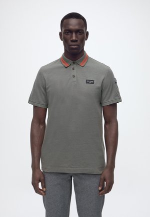 Uomo in piedi, rivolto verso di noi, che indossa una polo verde oliva con colletto a righe arancioni e pantaloni grigi su uno sfondo semplice.