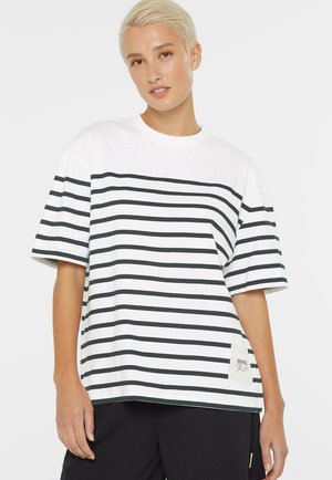 Person mit kurzen blonden Haaren, trägt ein weißes kurzärmeliges Hemd mit schwarzen horizontalen Streifen und schwarze Hose, steht vor weißem Hintergrund.