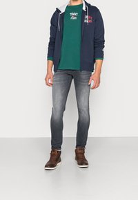 Laivastonsininen vetoketjuhuppari, jossa on harmaa vuori, vihreä pyöreäkauluksinen t-paita, jossa on valkoista tekstiä, harmaat slim-fit farkut ja ruskeat nauhakenkäbootit.