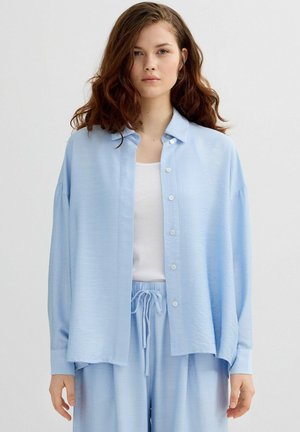 Joven mujer pelirroja que lleva una camisa de botones azul claro y holgada sobre un top blanco y pantalones azul claro a juego con cordón en la cintura.