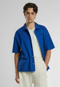 Blå kortærmet skjorte med knapper i bomuld med spidse kraver, båret over en hvid t-shirt med rund hals, parret med teksturerede, lyse bukser.
