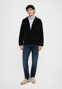 Black corduroy jacket with a button front, two front pockets, worn over a light blue shirt and paired with dark jeans and beige loafers.