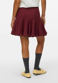 Burgunderfarbene, ausgestellte Rock mit einer weichen Textur, der am Saum Falten aufweist. Kombiniert mit grauen knielangen Socken und gelben Ballerinas.