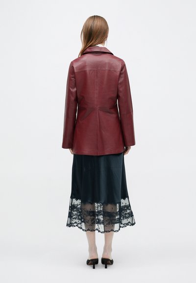 Γυναίκα με μπορντό δερμάτινο μπουφάν, μαύρη φούστα μίντι με διάφανο δαντελένιο τελείωμα και μαύρα ψηλοτάκουνα mules, όρθια με την πλάτη προς τα εμπρός σε λευκό φόντο.