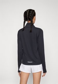 Czarna sportowa koszulka z długim rękawem i wysokim kołnierzem, gładka w dotyku, z białym akcentem na brzegu. Noszona z białymi szortami.