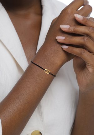 Eine Nahaufnahme einer Hand mit manikürten Nägeln, die ein schwarzes Armband mit einem goldenen Unendlichkeitssymbol trägt, vor einem hellen Hemd.