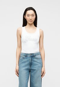 Femme aux cheveux longs et foncés portant un haut blanc sans manches avec le logo "Guess" et un jean taille haute bleu clair, debout devant un fond uni.