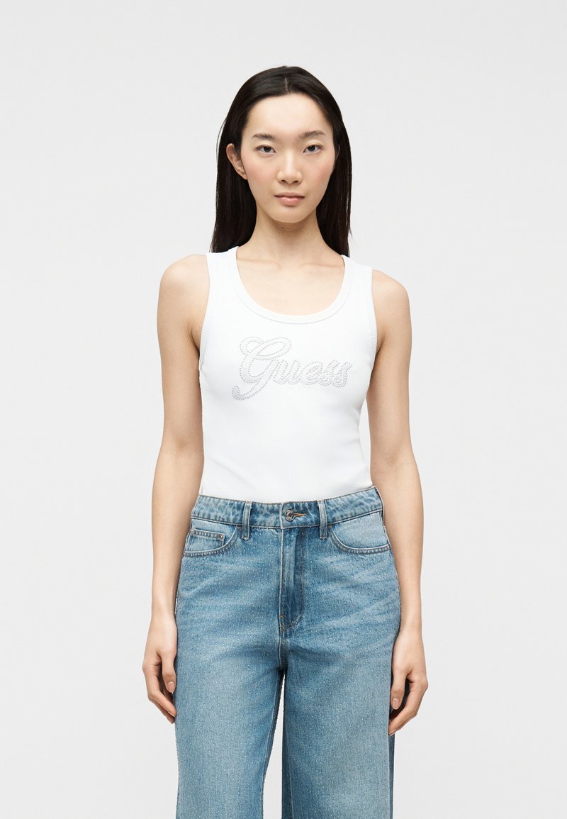 Femme aux cheveux longs et foncés portant un haut blanc sans manches avec le logo "Guess" et un jean taille haute bleu clair, debout devant un fond uni.