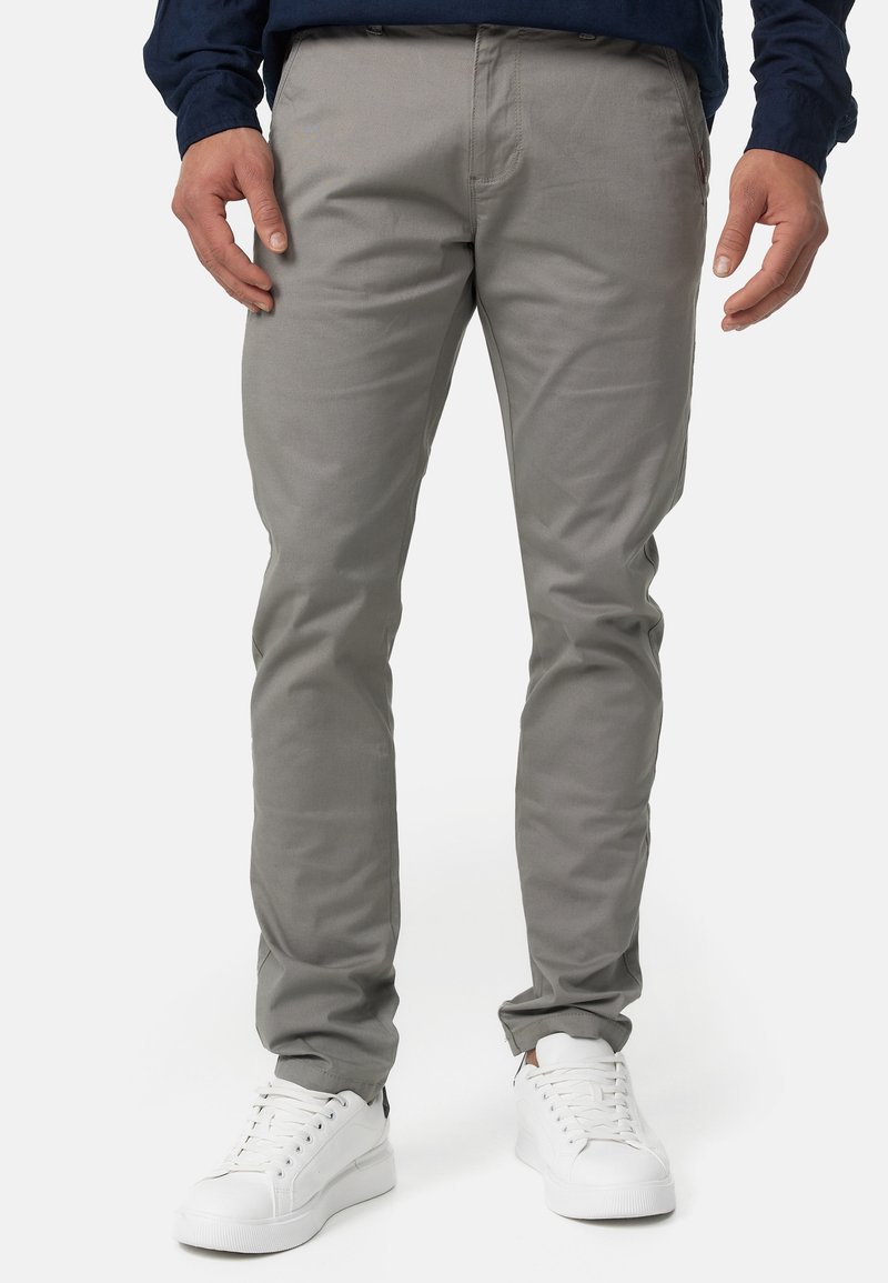 Homme portant un pantalon slim gris clair et des baskets blanches, debout les mains détendues le long du corps sur un fond uni.