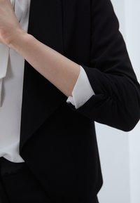 Schwarze Jacke aus weichem Stoff mit leichtem Drapieren, dazu eine weiße Bluse mit einem strukturierten Kragen, der aus den hochgekrempelten Ärmeln hervorschaut.
