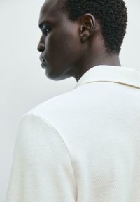 Blazer bianco con texture e finitura morbida, caratterizzato da un colletto a punte e dettagli di cucitura visibili, indossato da un modello visto di profilo.
