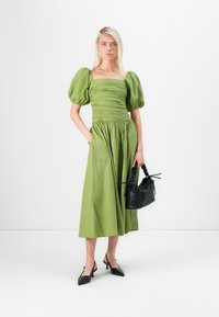 Robe verte mi-longue avec des manches bouffantes et une taille froncée ; associée à un sac à main en cuir texturé noir et des escarpins noirs à bout pointu.