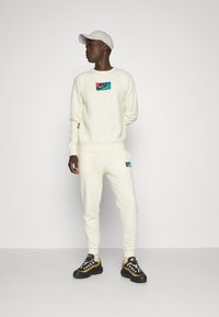 Cremefarvede sweatshirts og sweatpants sæt med et farverigt Nike-logo patch, kompletteret med en lys cap og atletiske sko med sort sål.